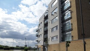 Un immeuble résidentiel à plusieurs étages, doté de balcons, d’une allée pavée et d’un espace verdoyant agrémenté de petits arbres.
