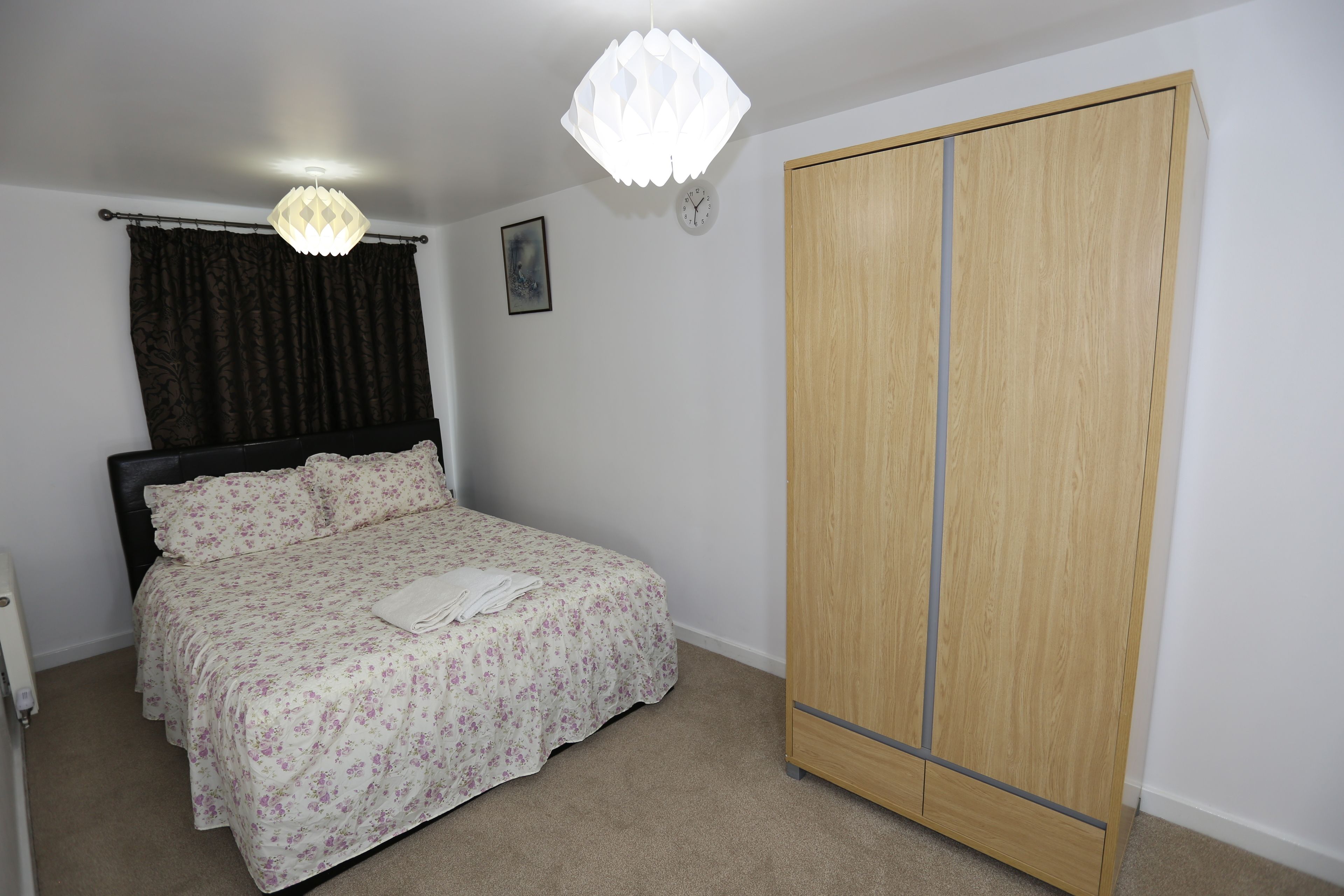 A bedroom with a bed, a wardrobe, a hanging light fixture, and a clock on the wall.