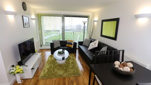 A modern living room with a flat-screen TV, a sofa set, a dining table, and a large window with blinds. - 2 Beds Riverview Tideslea Apartment (London)