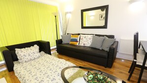 A modern living room with a black sofa, a floral bedspread, a glass coffee table with a bowl of fruit, and a bright green wall. - 2 Beds Riverview Tideslea Apartment (London)