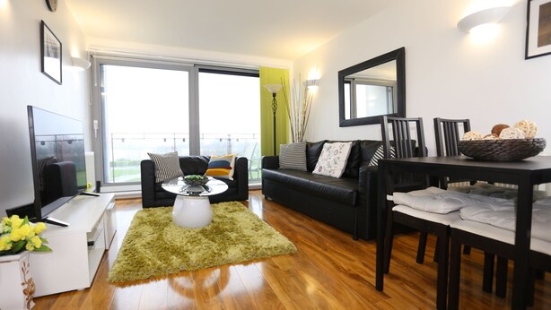 A modern living room with a sofa, coffee table, and dining table. There is a TV, a plant, and a window with a view. - 2 Beds Riverview Tideslea Apartment (London)