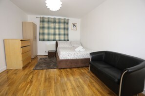 A bedroom with a bed, a black leather sofa, a wooden wardrobe, a plaid curtain, and a radiator.