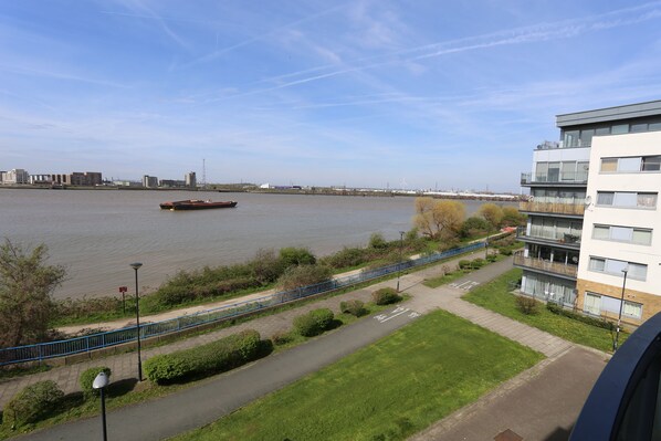 A riverside walkway with a blue fence, a grassy area, and a building with balconies.