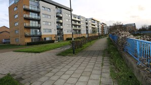 A multi-story residential building with balconies, a paved walkway, and a blue fence. - 2 Beds Riverview HillHouse Apartment (London)