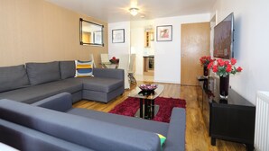 A modern living room with a grey sectional sofa, a small black table with a bowl, and a red rug. There is a flat-screen TV mounted on the wall and a wooden door leading to another room. - 2 Beds Riverview HillHouse Apartment (London)