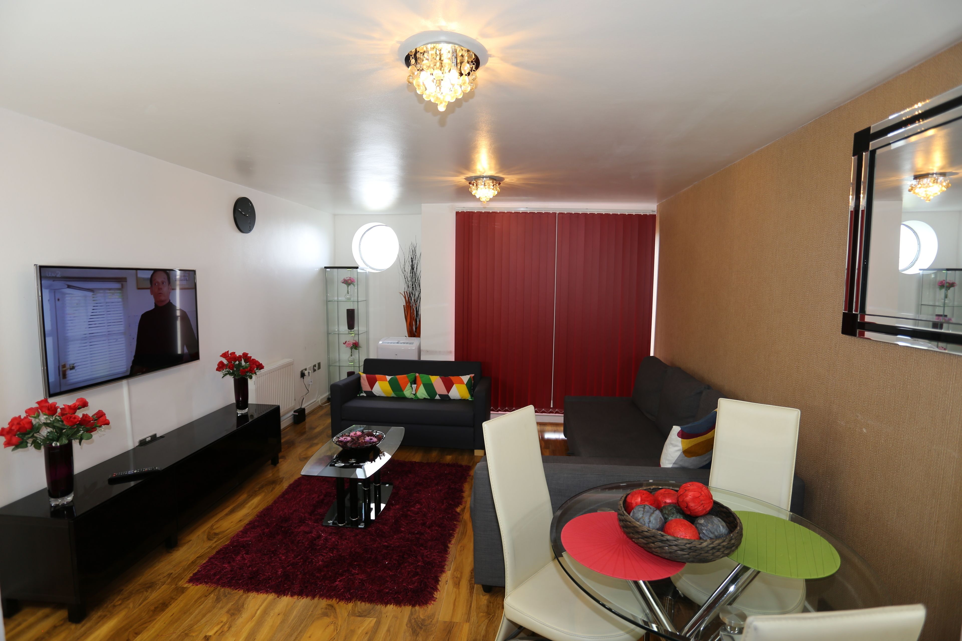 A modern living room with a flat-screen TV, a sofa, a dining table with chairs, and a red curtain.