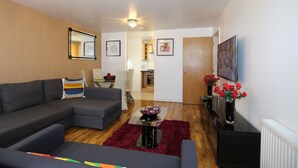 A modern living room with a sectional sofa, a coffee table, and a red rug. There is a kitchen in the background and a door leading to another room. - 2 Beds Riverview HillHouse Apartment (London)