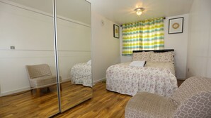 A bedroom with a large mirror, wooden flooring, a bed with floral bedding, a chair, and a window with striped curtains. - 2 Beds Riverview HillHouse Apartment (London)