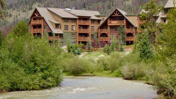 A lodge with multiple wooden structures, a river in the foreground, and a forested mountain backdrop. - Walk to Mountain House, Great Slope Views, Private Laundry, Shared Hot Tubs (Keystone)