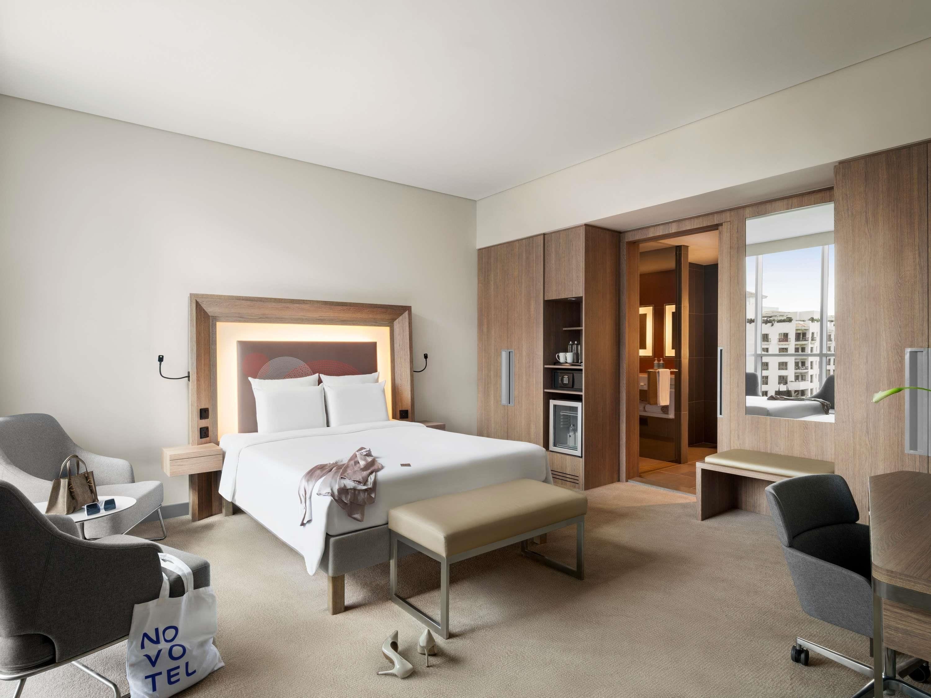 A hotel room with a bed, a desk, a chair, a bench, a wardrobe, and a view of the city through a window.