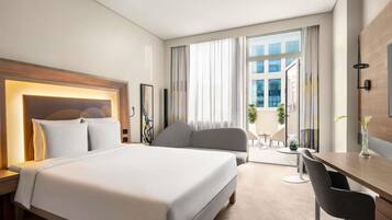 Quarto de hotel com uma cama grande, uma escrivaninha, uma cadeira e vista da cidade por grandes janelas.