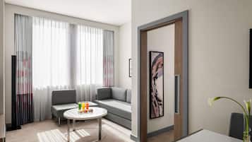 Quarto de hotel moderno com um sofá cinzento, uma mesa de centro redonda com petiscos, uma janela grande com cortinas leves e uma obra de arte minimalista na parede.