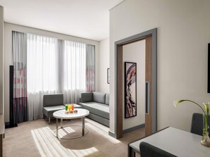 A modern hotel room with a grey sofa, a round coffee table with snacks, a large window with sheer curtains, and a minimalist artwork on the wall. - Novotel Bur Dubai (Dubai)