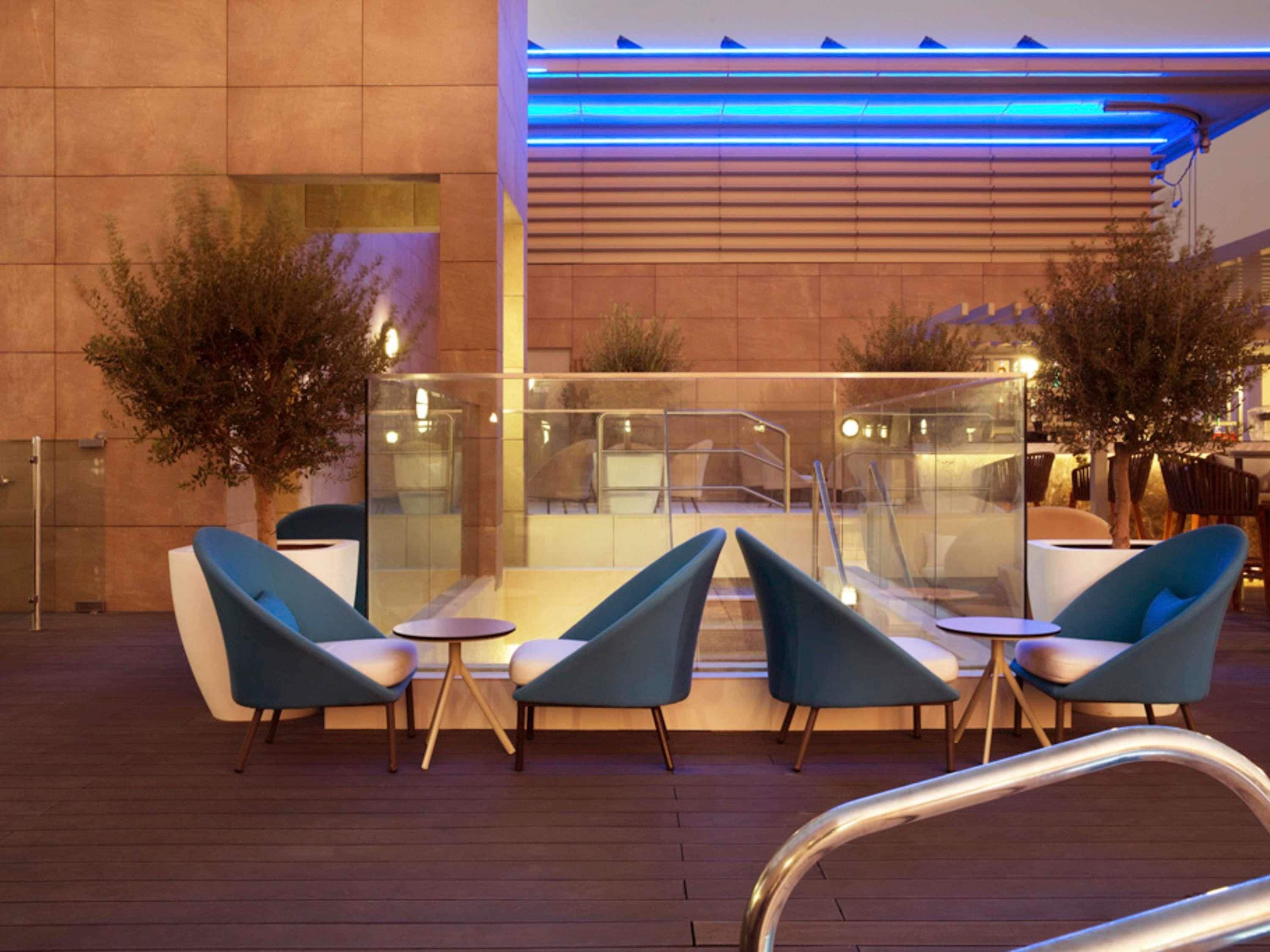 Un área de estar al aire libre con sillas y mesas modernas, un separador de vidrio y un edificio con iluminación azul al fondo.