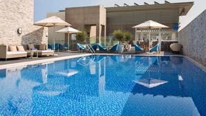 A modern pool area with lounge chairs and umbrellas, a bar, and a stone wall. - Novotel Bur Dubai (Dubai)