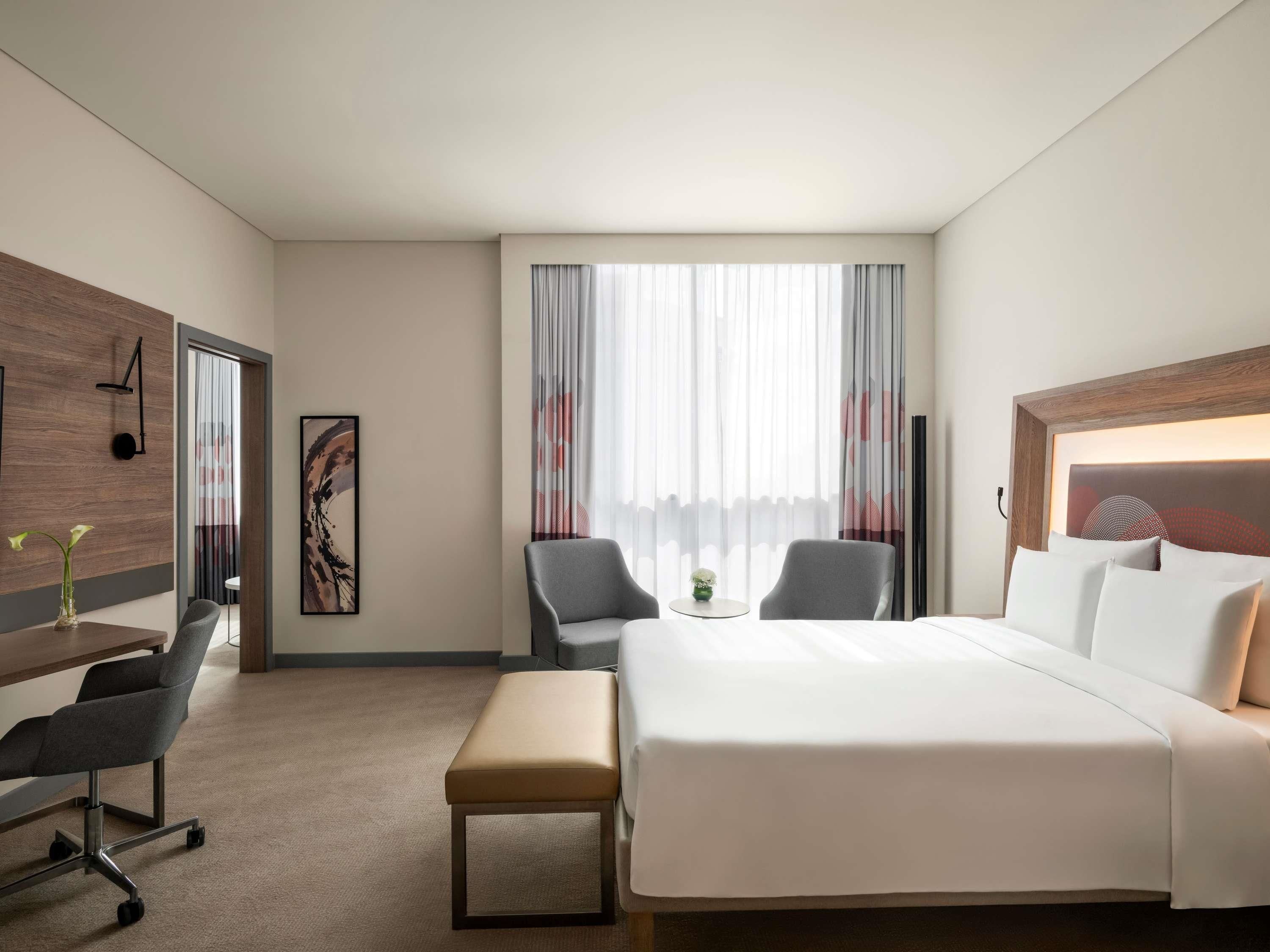 A hotel room with a bed, two armchairs, a desk with a chair, a small table, and a large window with curtains.