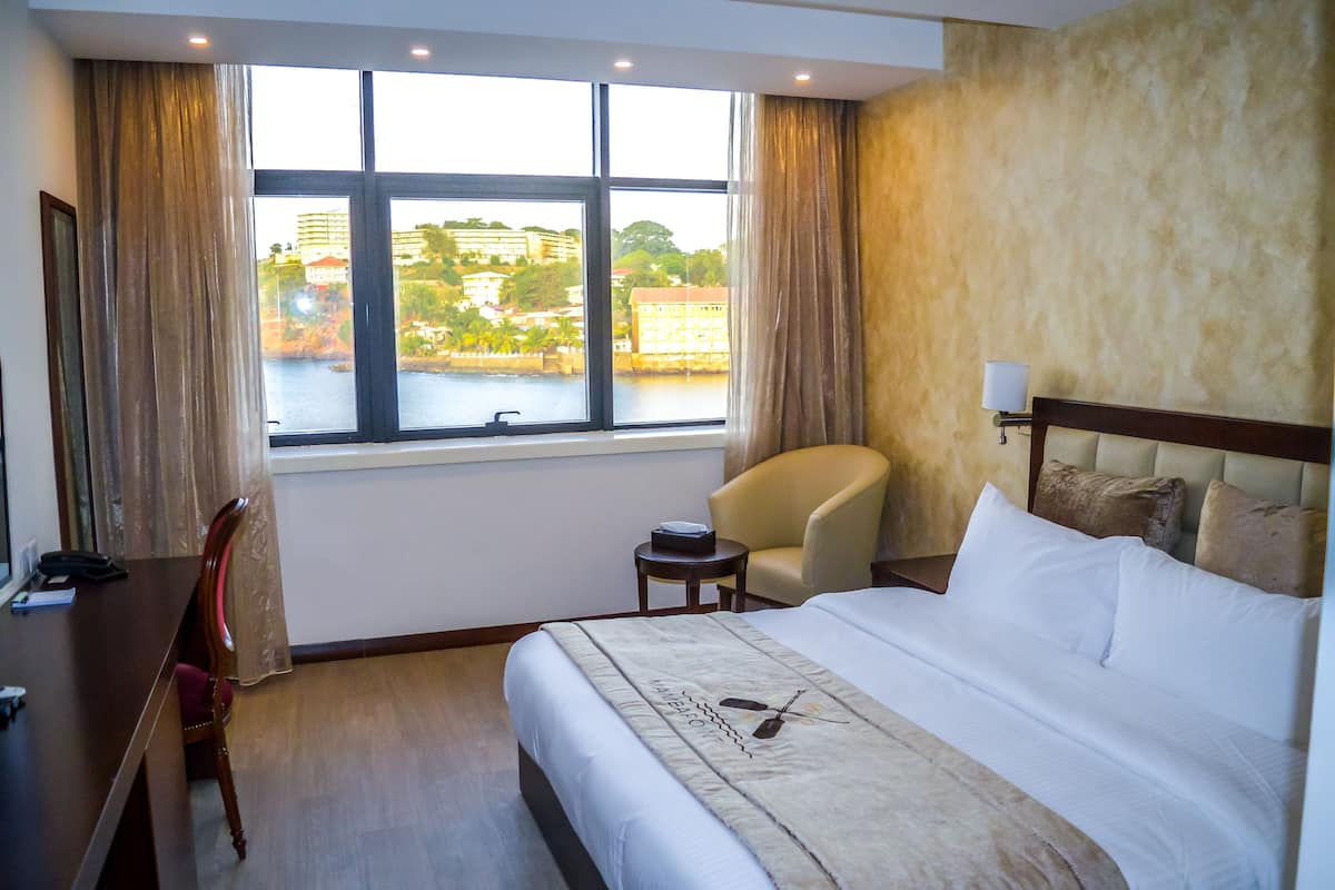 A hotel room with a large bed, a desk, a chair, a small table, and a view of a body of water through a window.