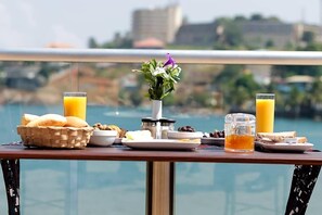 A breakfast table with various food items and drinks, a vase with flowers, and a view of a body of water and buildings in the background. - Mamba Point Hotel Freetown (Freetown)