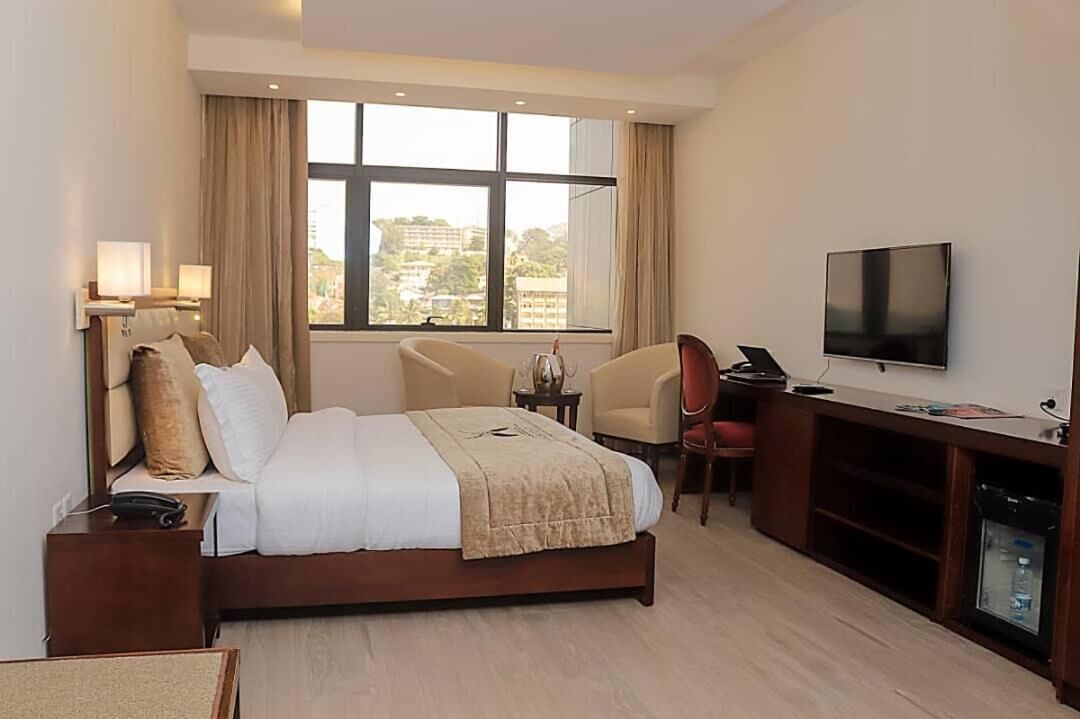 A hotel room with a bed, bedside lamps, a TV, a desk, and a view of buildings outside the window.