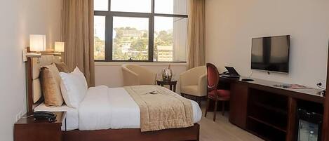 A hotel room with a bed, bedside lamps, a TV, a desk, and a view of buildings outside the window.