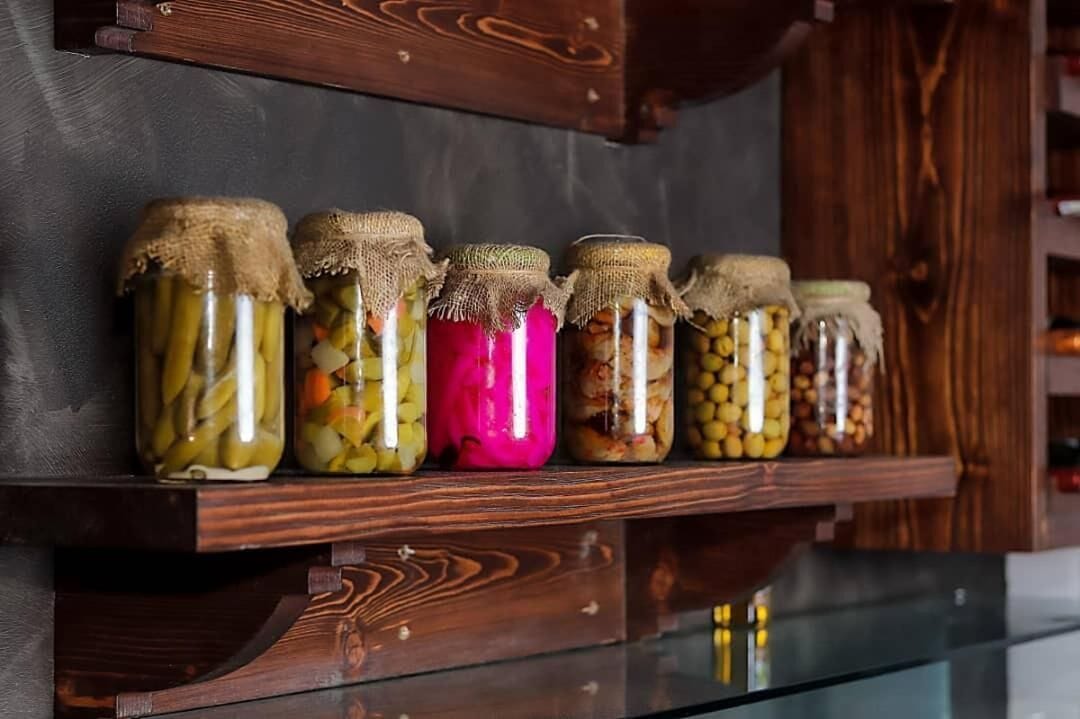A wooden shelf with six glass jars of pickled vegetables.