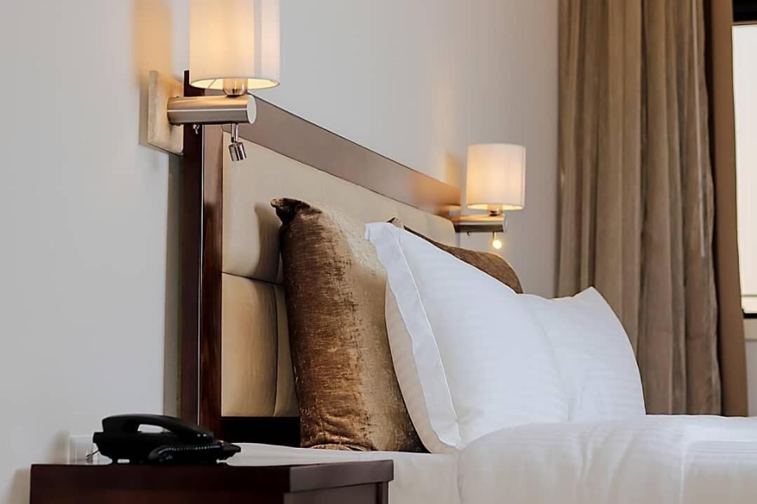 A hotel room with a bed, two wall-mounted lamps, a telephone, and a window with curtains.