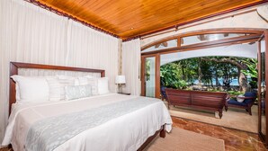 A spacious bedroom with a large bed, a wooden ceiling, and a glass door leading to an outdoor seating area. - Blue Jewel Oceanfront Private Villa (Tamarindo)