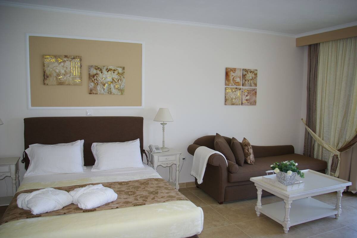 A hotel room with a bed, a brown sofa, a white coffee table with a flower vase, and artwork on the walls.
