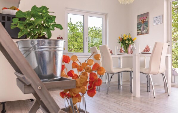 Un espace repas moderne avec une table et des chaises blanches, une plante en pot et une fenêtre donnant sur des arbres.