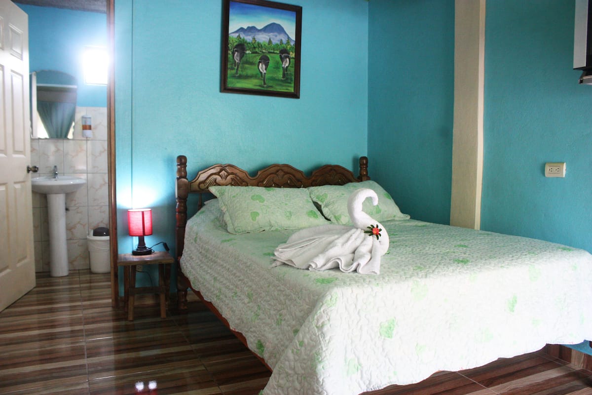 A bedroom with a bed, a nightstand, a lamp, a painting, and a bathroom with a sink and mirror.