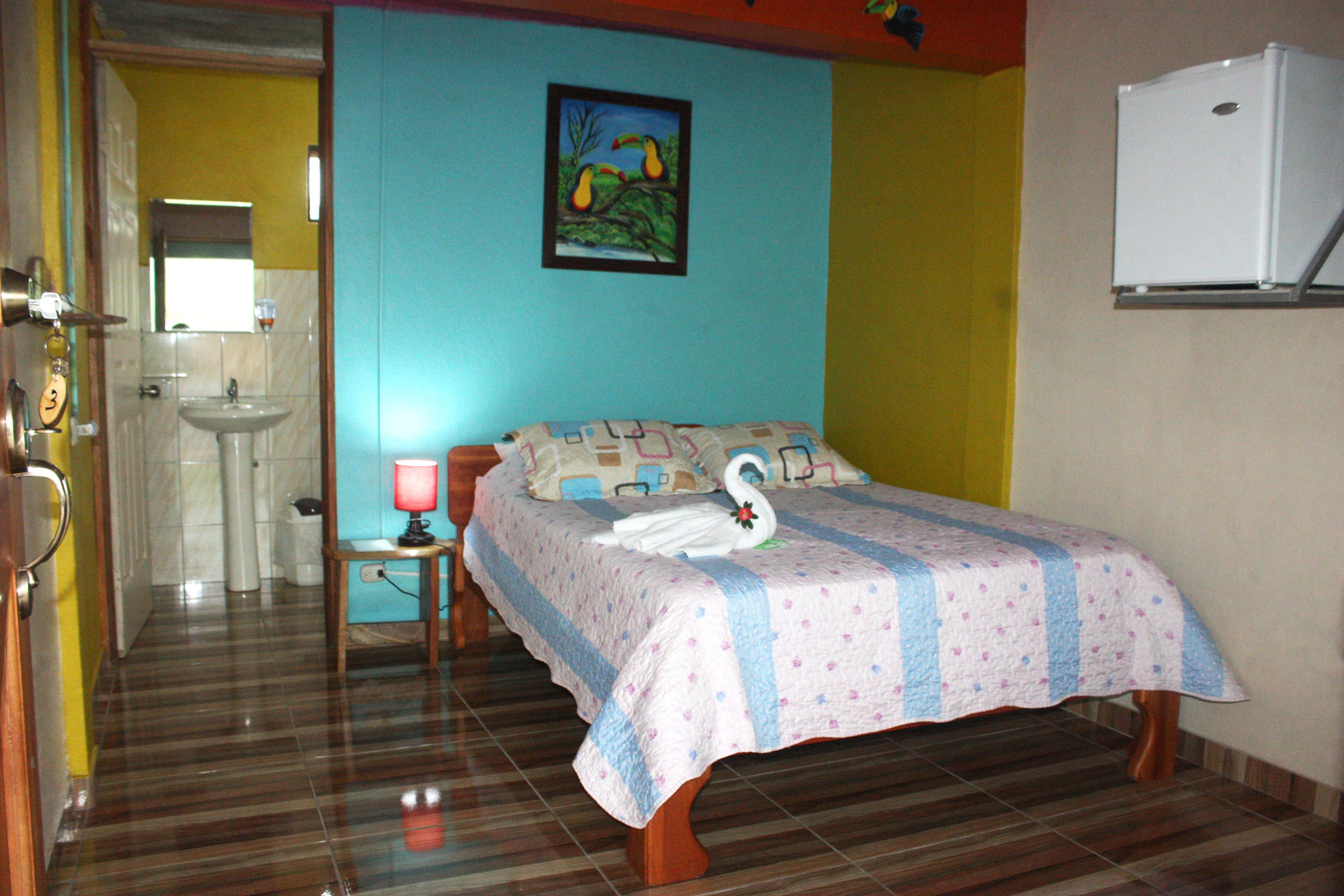 A bedroom with a bed, a nightstand, a lamp, a wall-mounted air conditioner, and a bathroom visible through an open door.