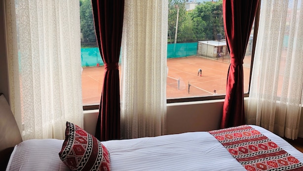 Habitación de hotel con cama, ventana con cortinas y vista a una cancha de tenis.