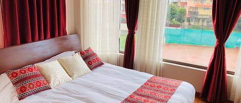 A neatly made bed with patterned pillows, a wooden headboard, and a view of a tennis court through sheer curtains.