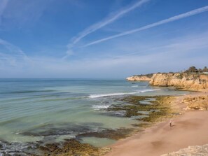 Un paysage côtier caractérisé par un ciel dégagé, une plage de sable, des affleurements rocheux et une mer calme.