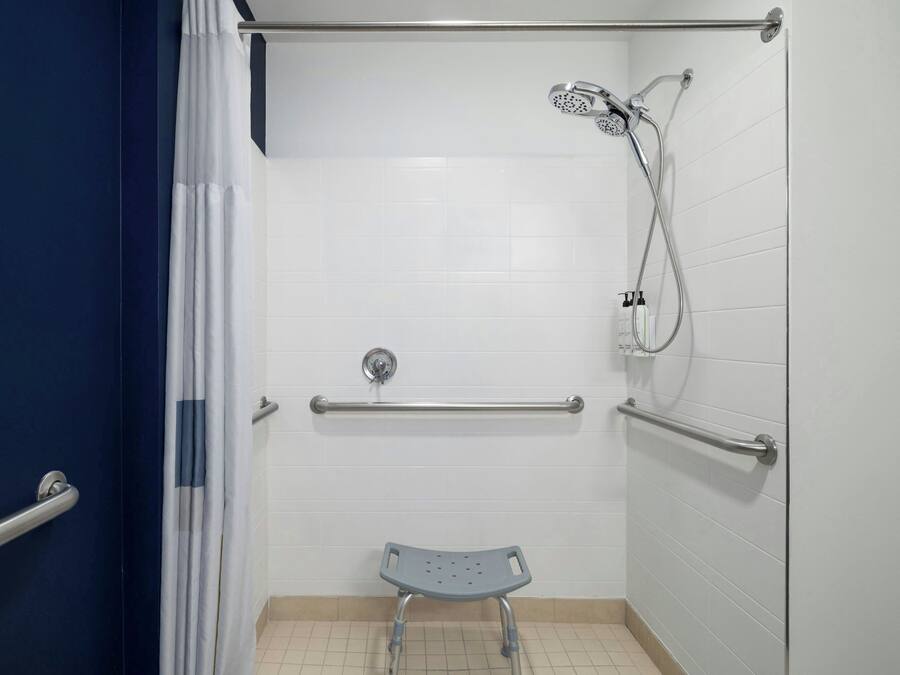 Une salle de bain avec des murs carrelés de blanc, une douche équipée d’un pommeau de douche mobile, une barre d’appui et un banc accessible aux personnes en fauteuil roulant.