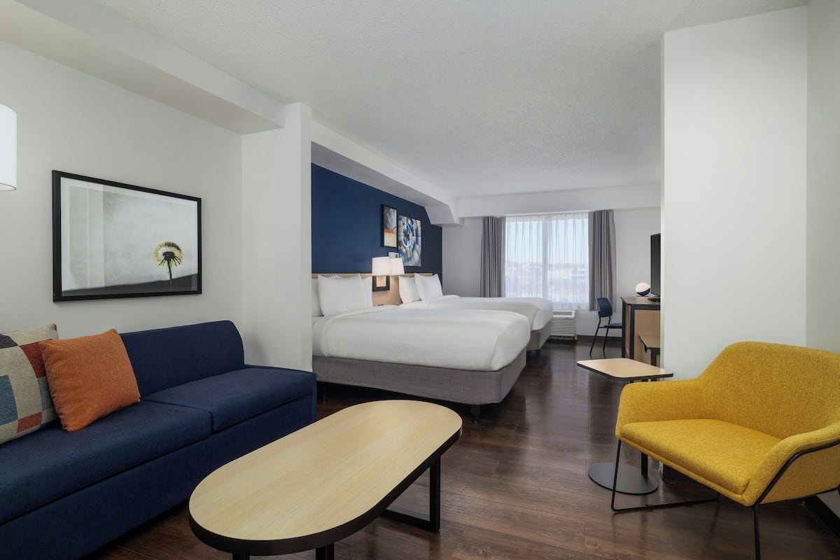 A hotel room with a blue sofa, a yellow armchair, a bed, a desk, and a TV.