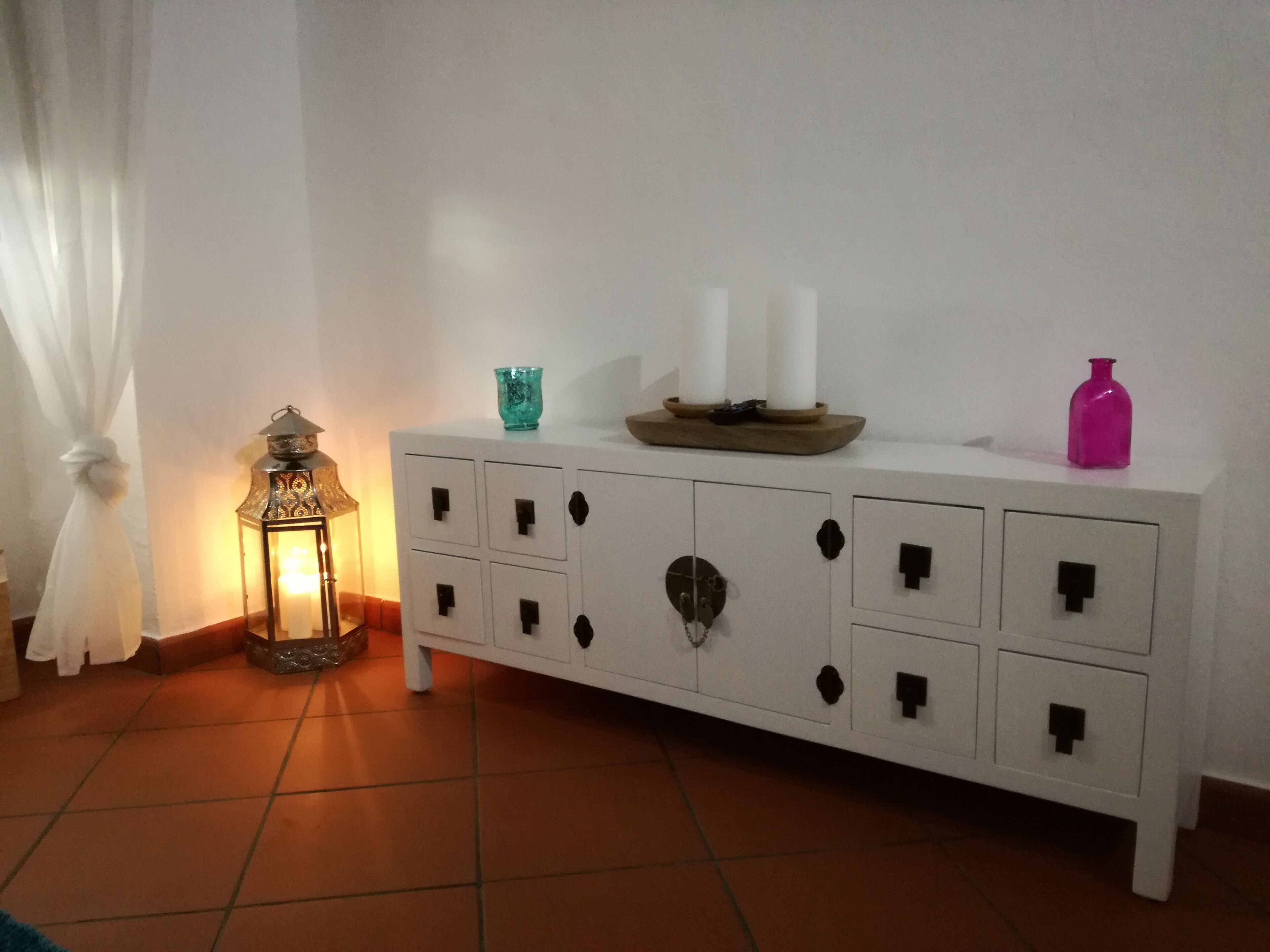 Un mueble blanco con varios cajones, un florero de vidrio color turquesa, dos velas blancas y una botella rosa encima.
