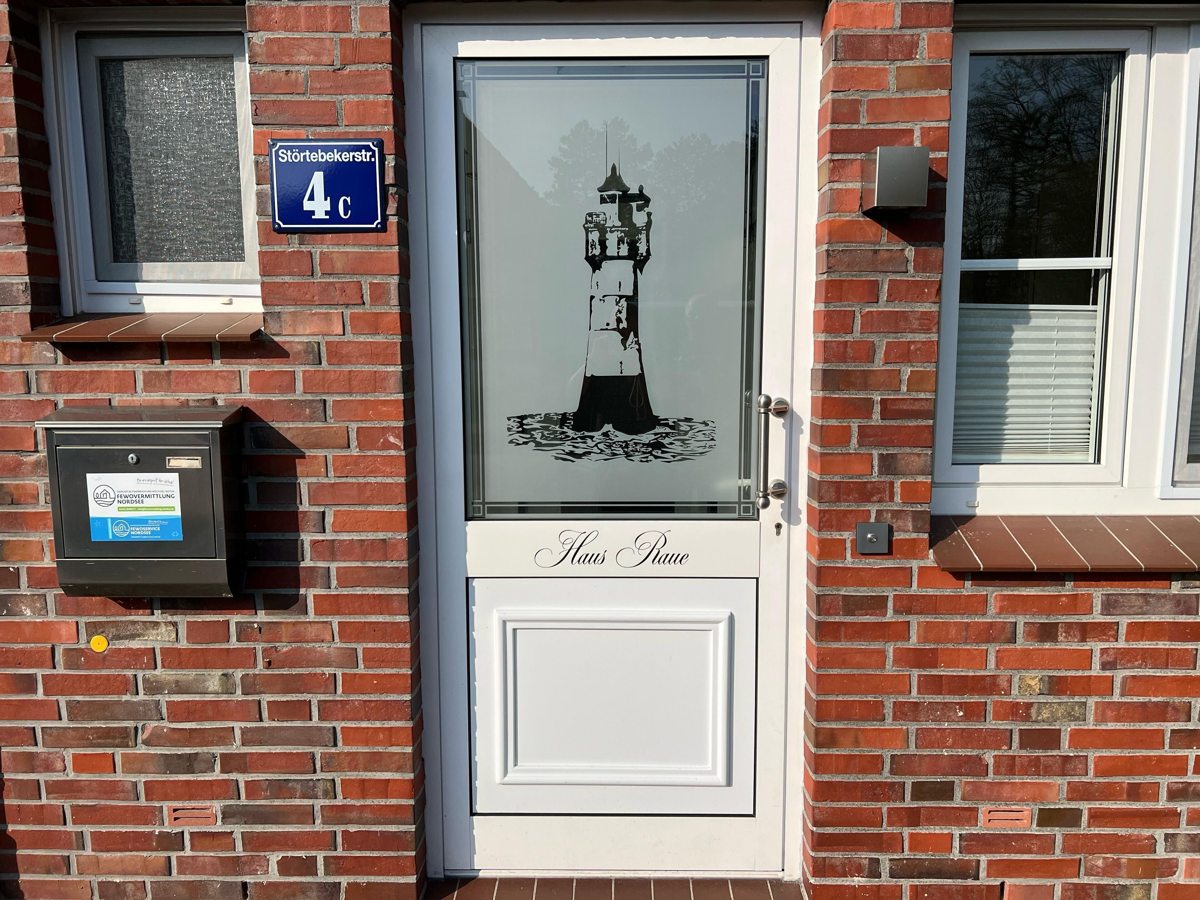 A white door with a glass panel featuring a lighthouse design, a blue street sign above the door, and a mailbox on the left.