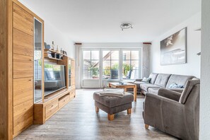 A modern living room with a wooden entertainment unit, a grey sofa, and a large window offering a view of the outdoors.