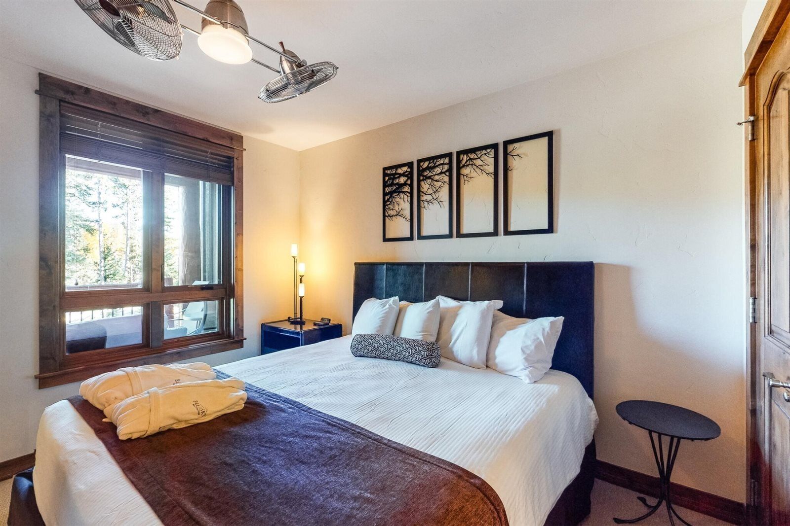 A neatly arranged bedroom with a large bed, a bedside table with a lamp, a small round table, and a window with blinds.