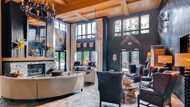 A spacious living room with a stone fireplace, leather sofas, and a large window overlooking a forest. - Luxury 3 Bedroom Ski in, Ski Out Mountain Vacation Rental Just Steps from the Snowflake Ski Lift (Breckenridge)