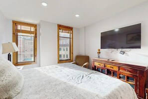 A hotel room with a bed, a desk, a chair, a TV, and two windows with blinds.