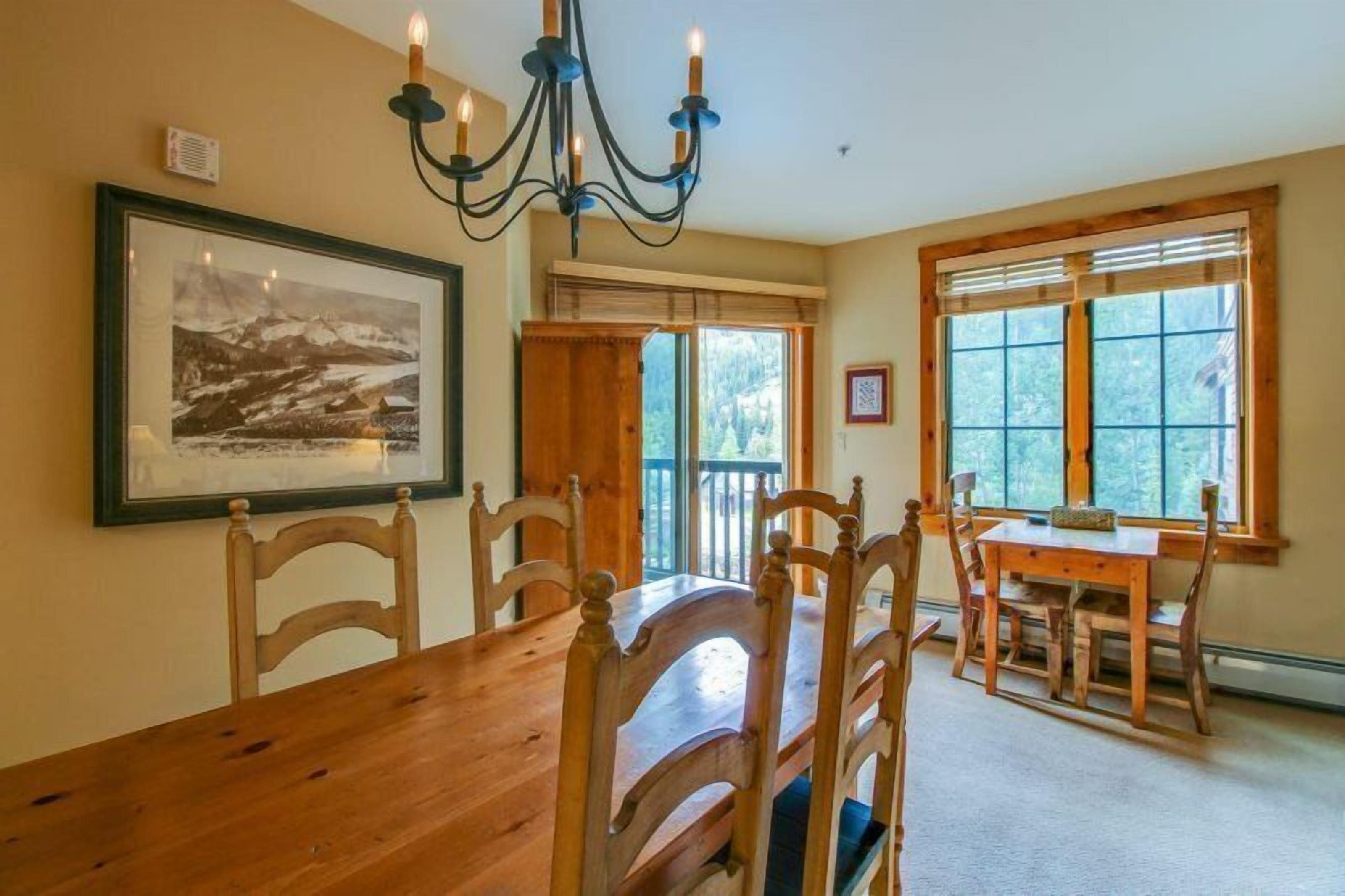 Une salle à manger avec une table et des chaises en bois, un lustre, un tableau encadré au mur et de grandes fenêtres donnant sur un paysage verdoyant.