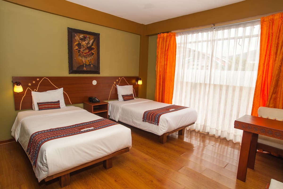 Ein Hotelzimmer mit zwei Betten, einem Holzkopfteil, einem Nachttisch, einem Schreibtisch und einem Fenster mit orangefarbenen Vorhängen.