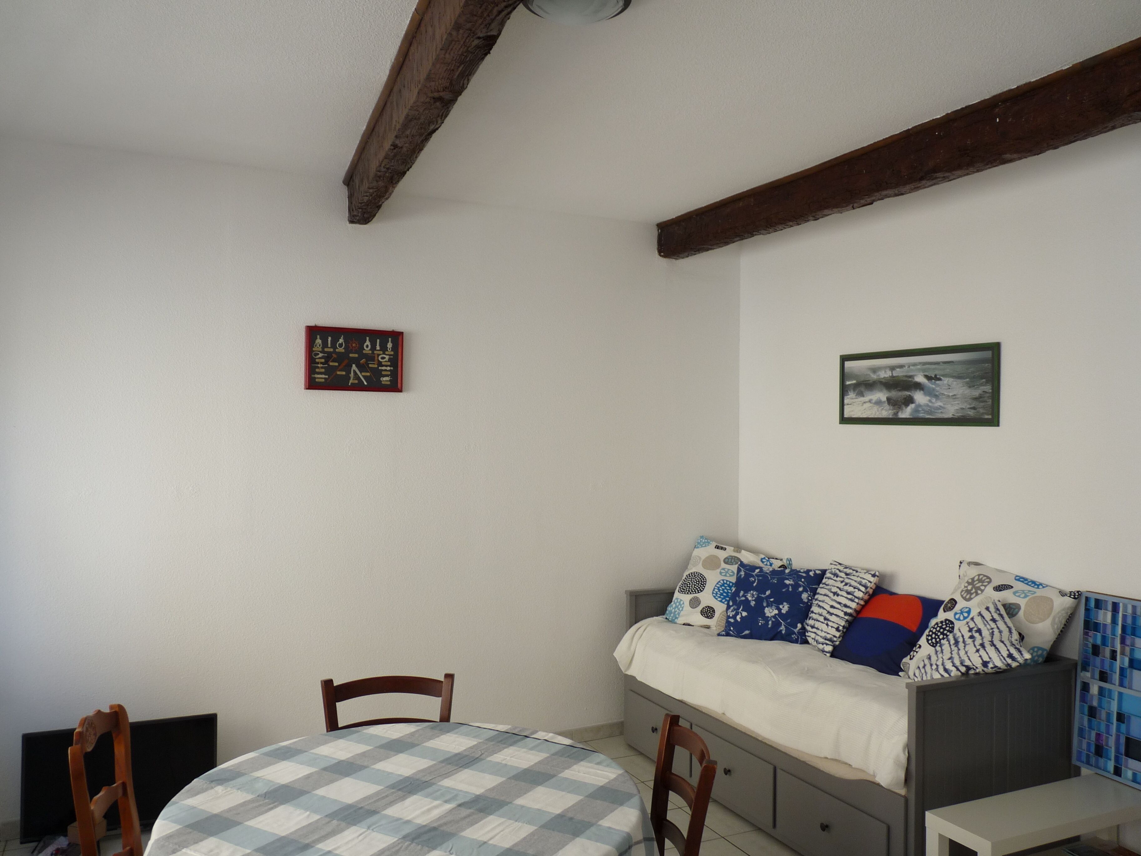 Una habitación con una cama, una mesa y una silla. Hay un cuadro en la pared y una pequeña mesa con mantel a cuadros.