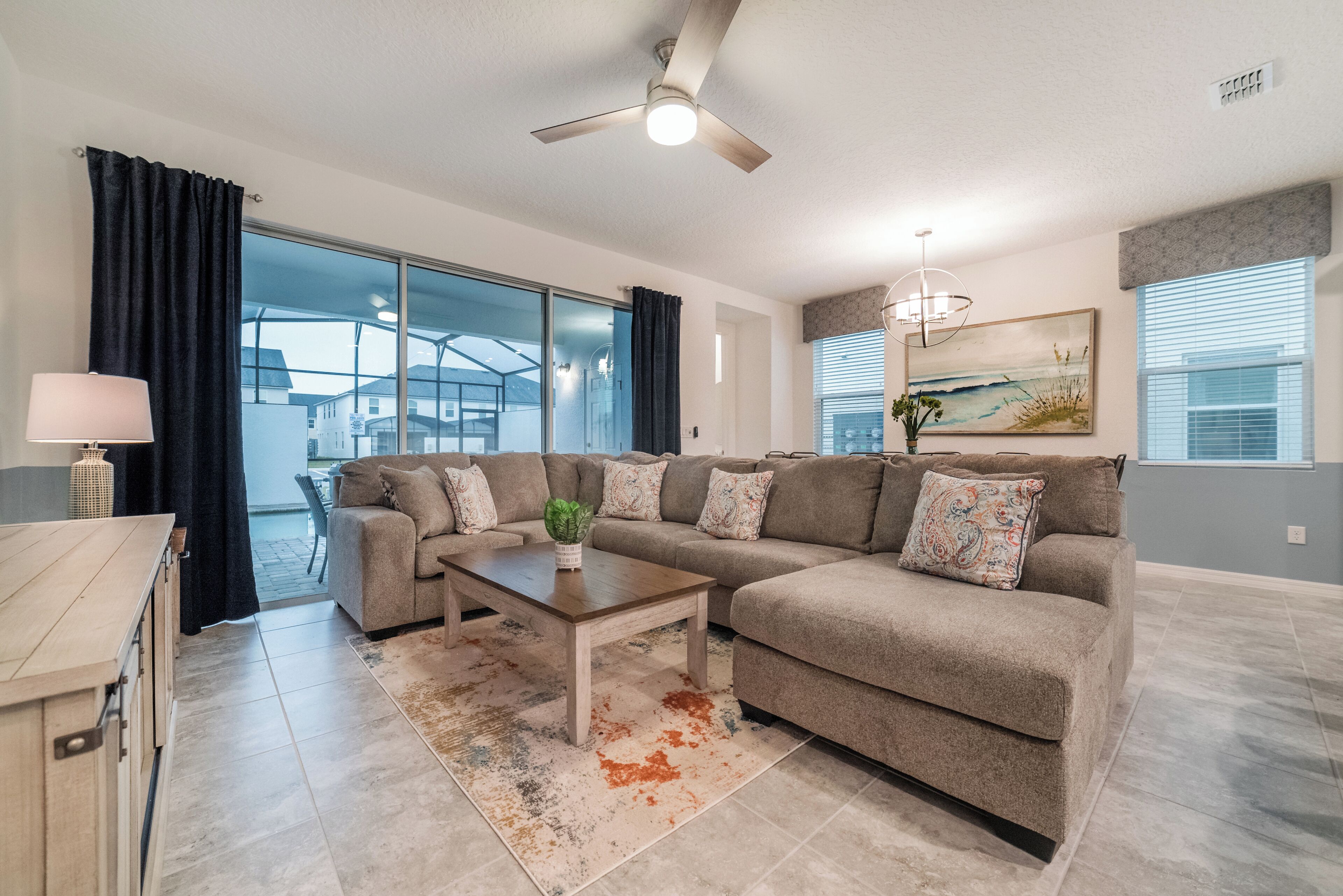 A modern living room with a sectional sofa, a coffee table, a ceiling fan, and large windows.