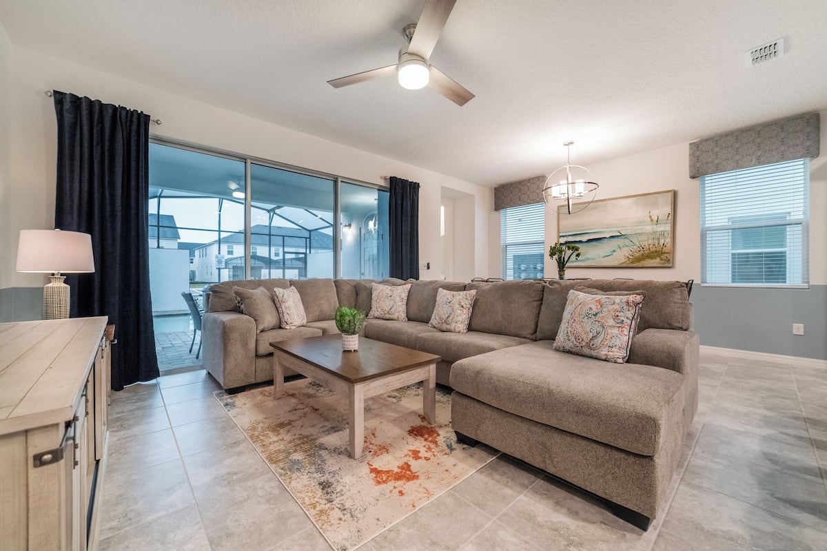 A modern living room with a sectional sofa, a coffee table, a ceiling fan, and large windows.