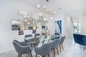 Uma sala de jantar moderna com mesa de vidro, cadeiras cinzentas, uma poltrona de veludo azul, um candelabro e uma cozinha ao fundo.