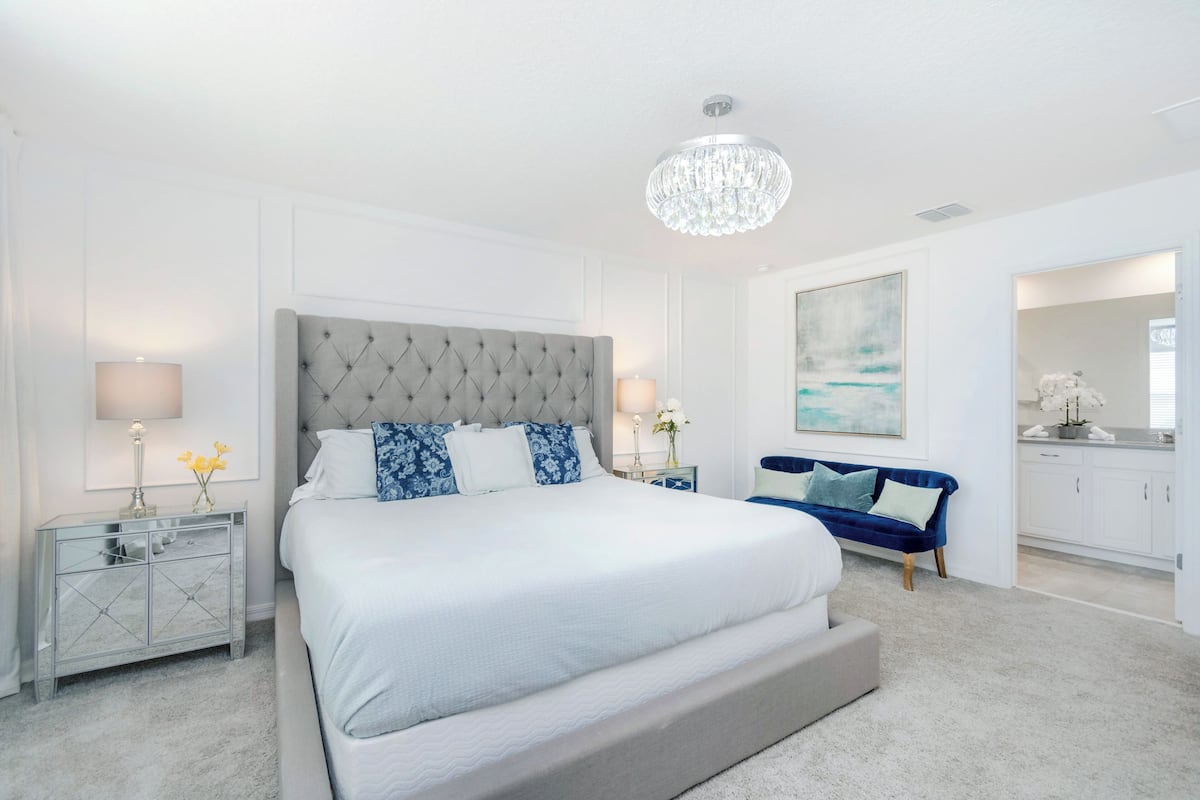 A bedroom with a large bed, two bedside tables, a chandelier, a blue sofa, and a painting on the wall.