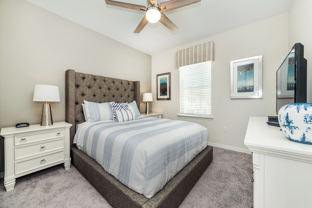 A bedroom with a tufted headboard, a bed with striped bedding, a nightstand with a lamp, a dresser, and a ceiling fan.